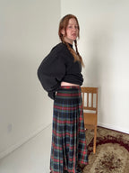 Person wearing a black sweatshirt and plaid skirt standing in a room with a chair and carpet.