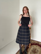 Person wearing a black top and plaid skirt in a room with a white wall and wooden chair.