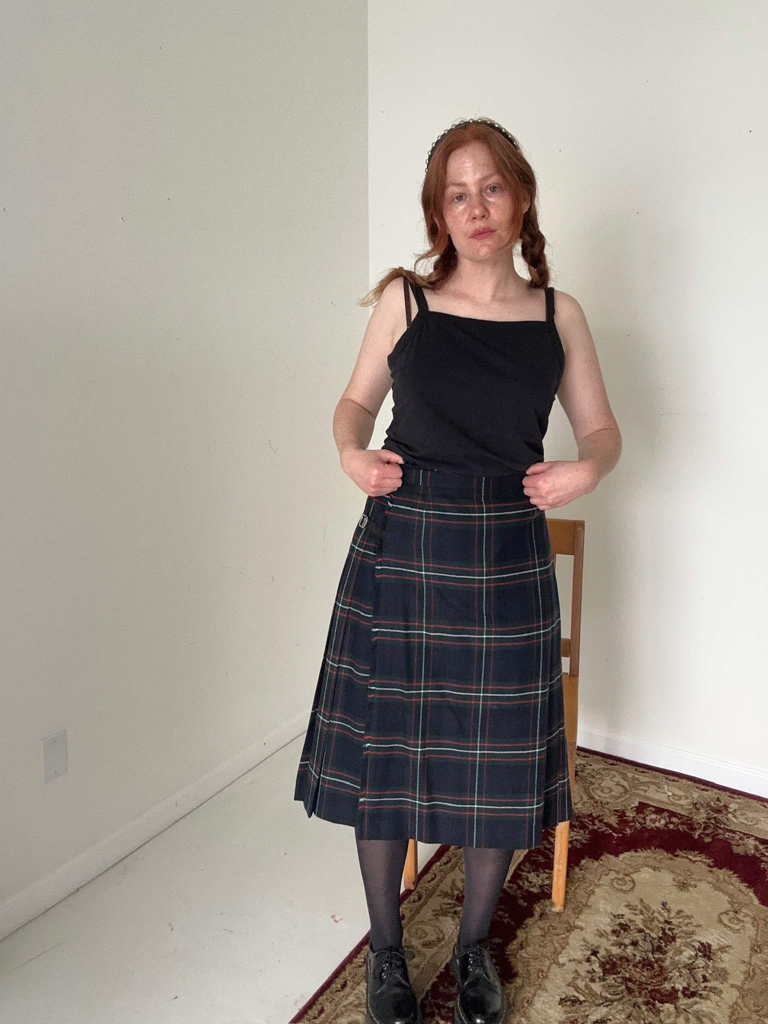 Person wearing a black top and plaid skirt in a room with a white wall and wooden chair.