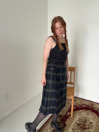 Person wearing a black tank top and plaid skirt standing in a room with a chair and carpet.