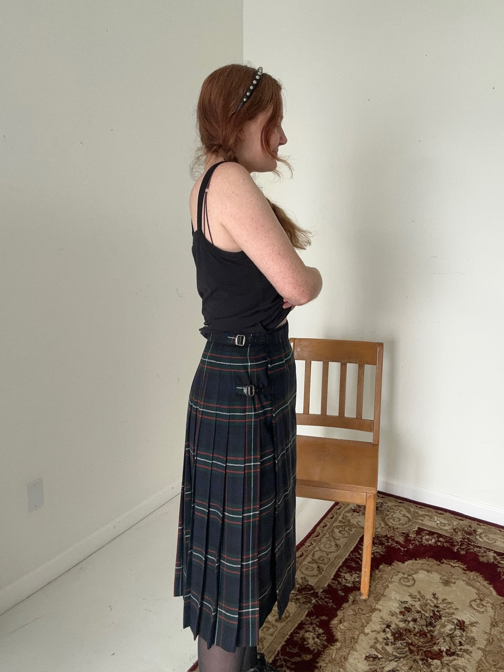 Person wearing a black tank top and plaid skirt standing in a room with a wooden chair and patterned rug.