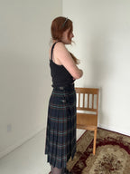 Person wearing a black tank top and plaid skirt standing in a room with a wooden chair and patterned rug.