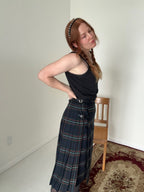 Person wearing a black tank top and plaid skirt standing in a room with a chair and carpet.