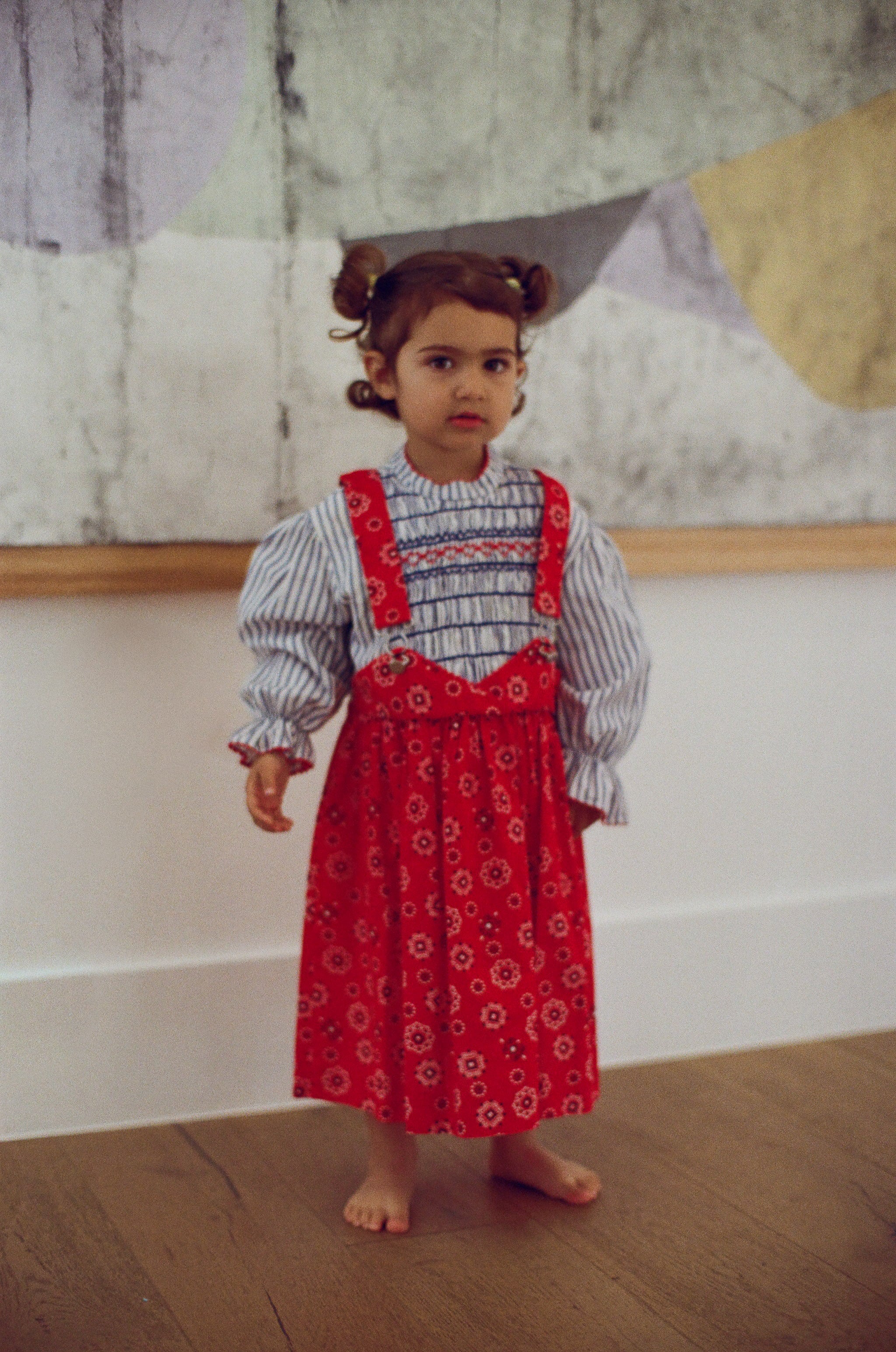 Child wearing a red patterned dress over a striped shirt against a textured wall.