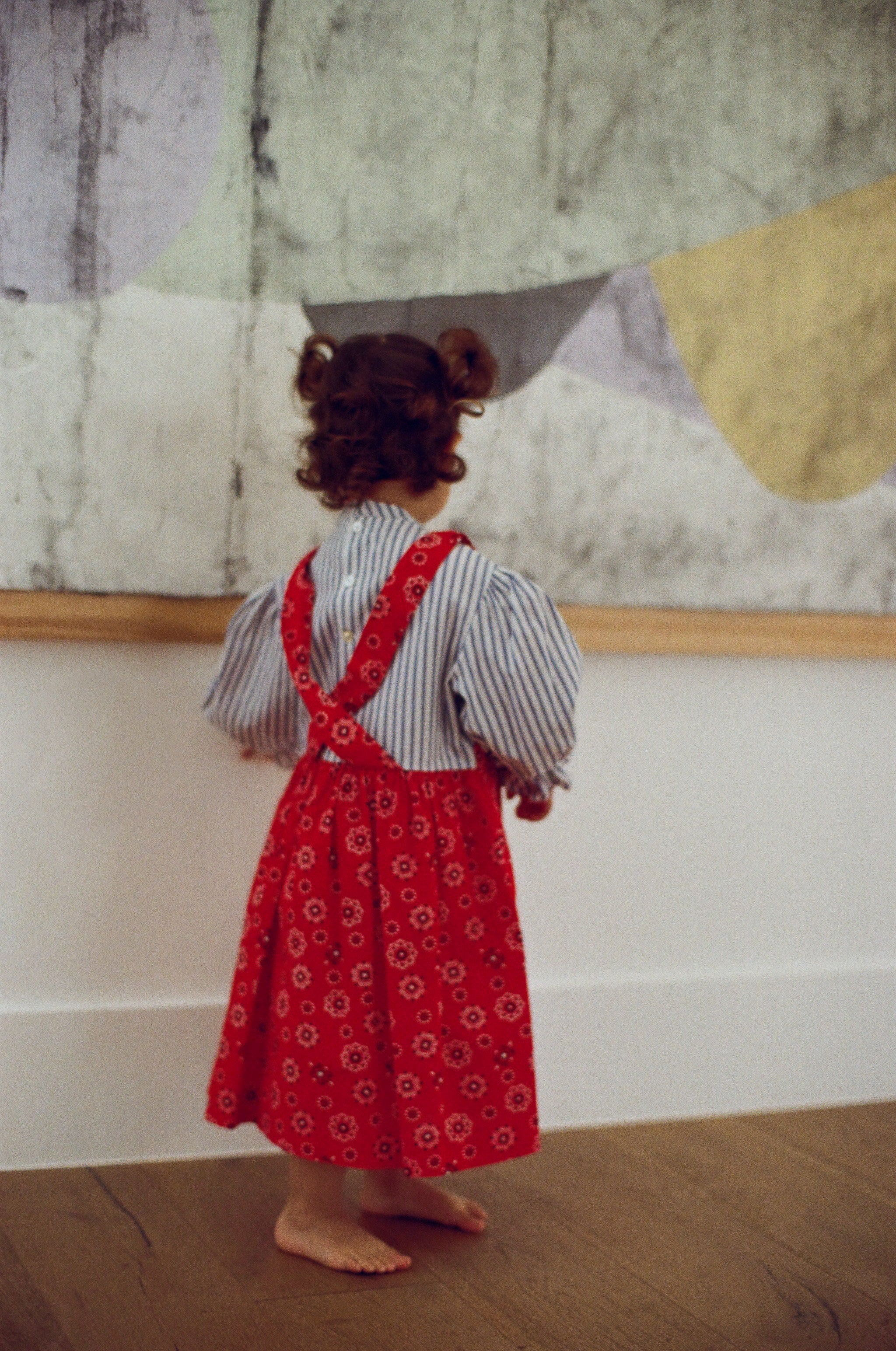Child wearing a red floral dress with suspenders standing in front of a wall with abstract art.