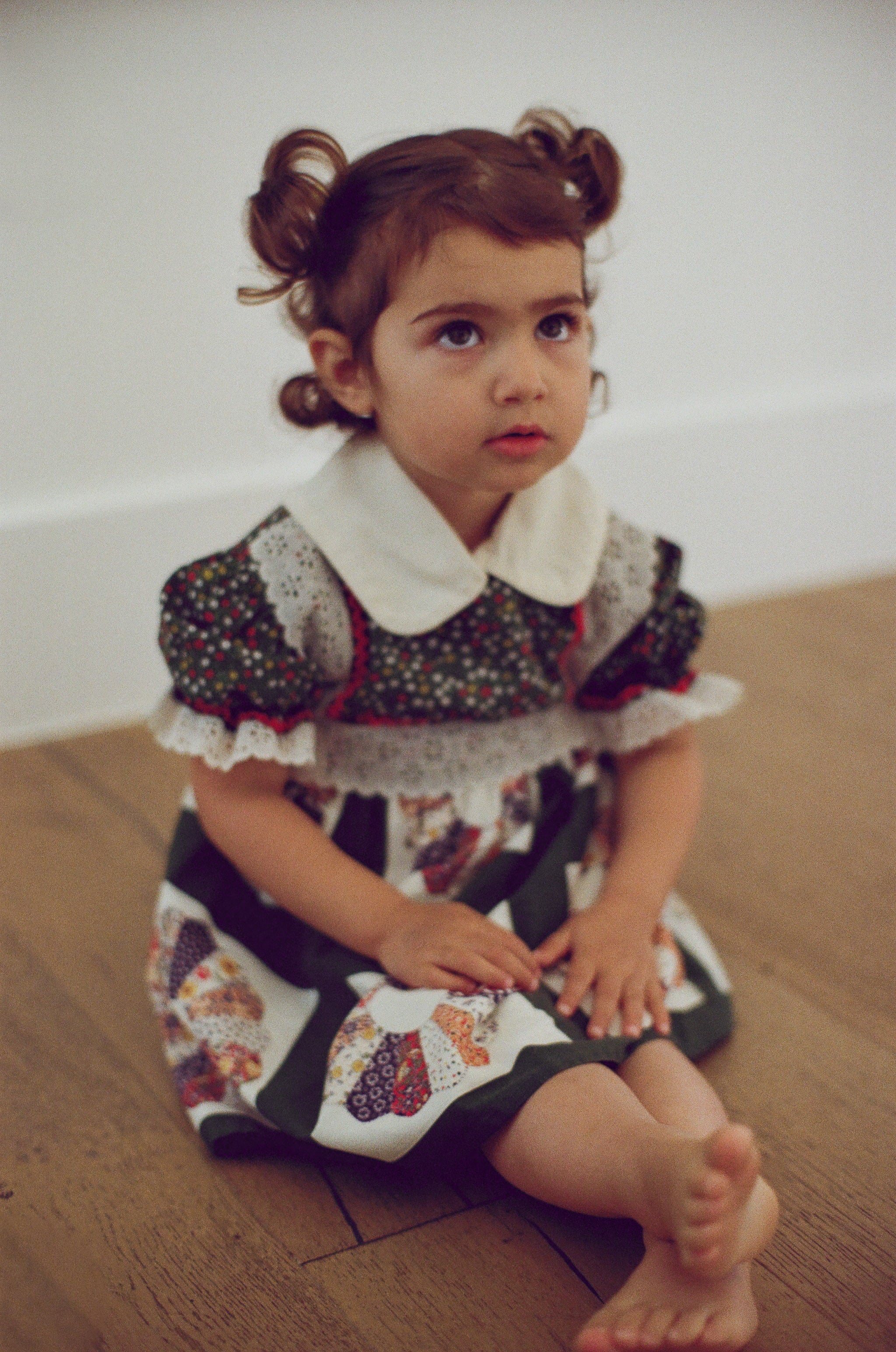 Young girl in a patterned dress sitting on a wooden floor.