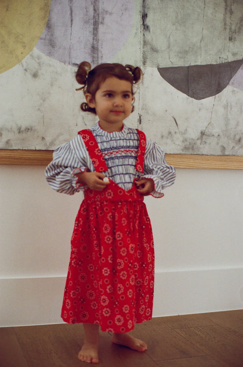 Young girl wearing a red dress with a patterned blouse against a textured wall.