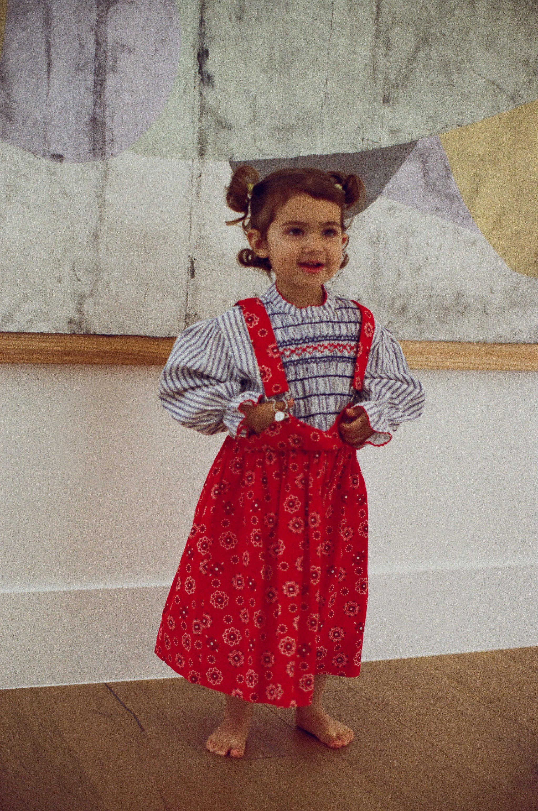 Young girl wearing a red patterned dress with suspenders standing indoors.