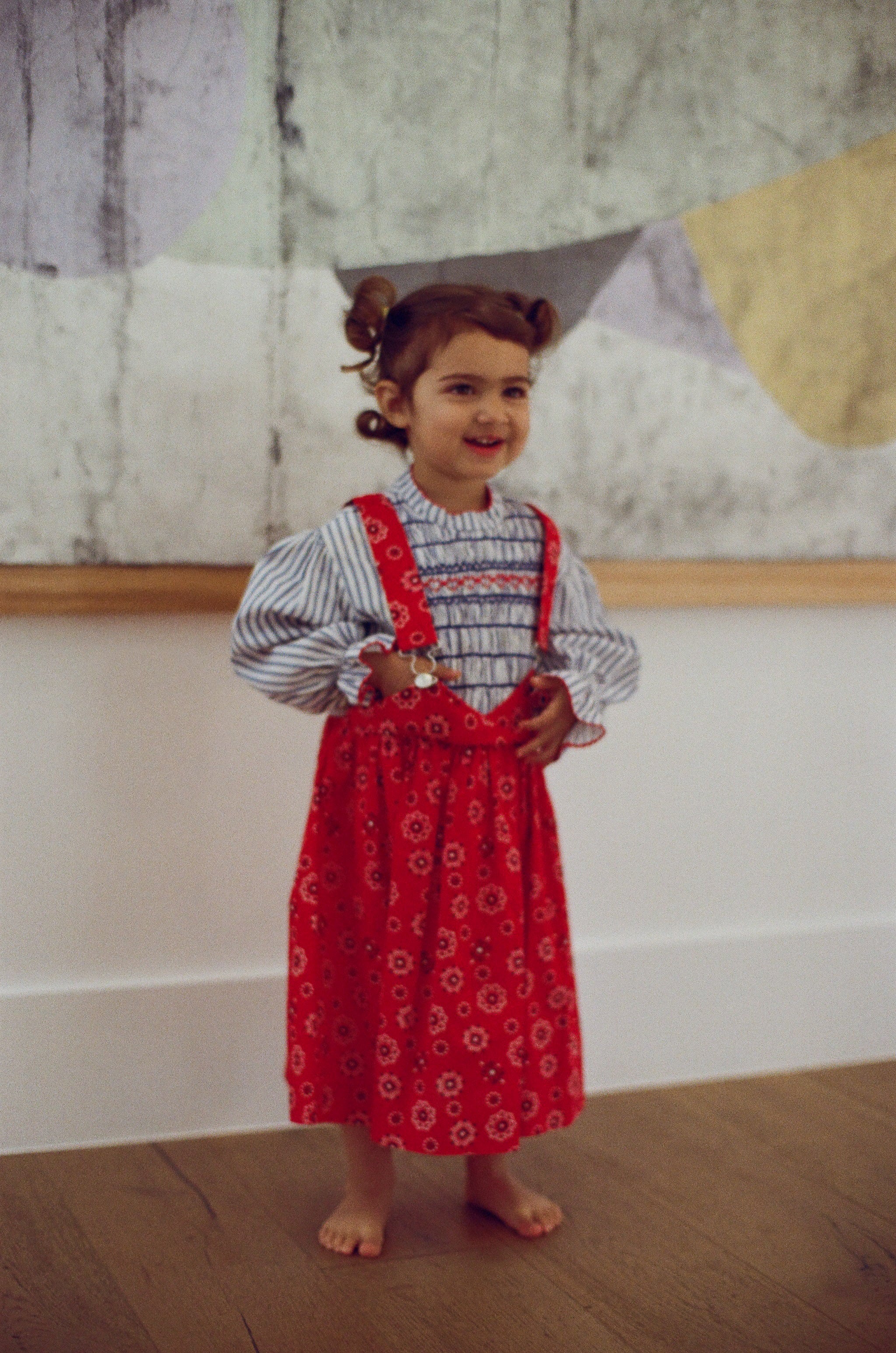 Child wearing a red patterned dress with suspenders standing indoors.