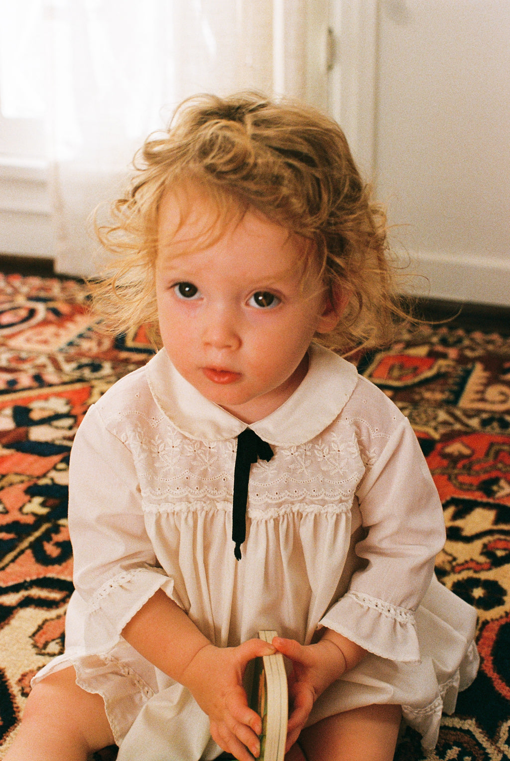 Young girl in a white dress sitting on a patterned rug.