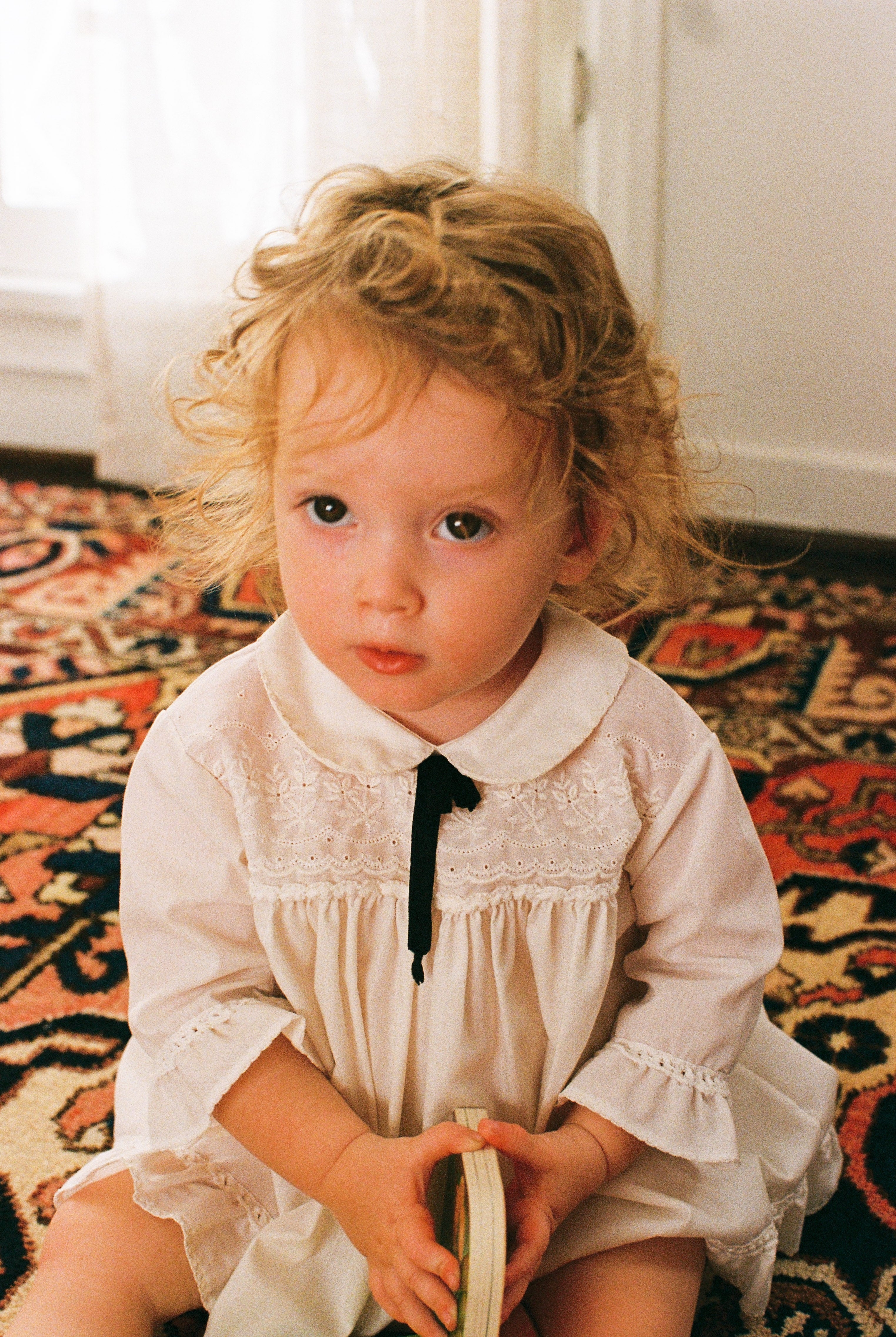 Young girl in a white dress sitting on a patterned rug.