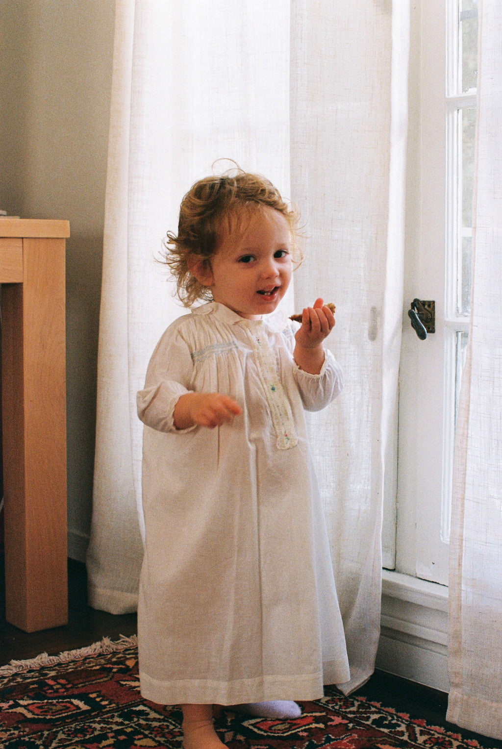 Child in a white dress standing in a room with white curtains and a wooden cabinet.