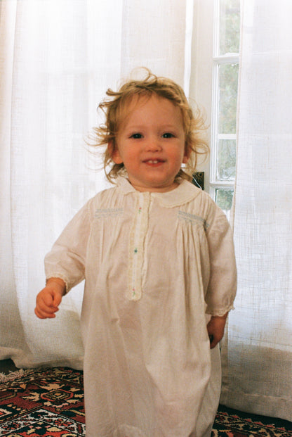 Child wearing a beige dress standing in front of white curtains