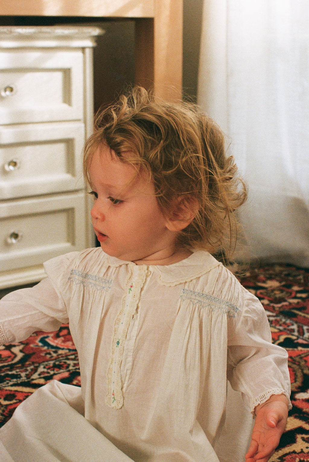 Child in a white dress sitting on a patterned rug with a neutral background