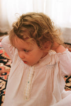 Child wearing a light pink dress with lace details, sitting on a patterned rug.