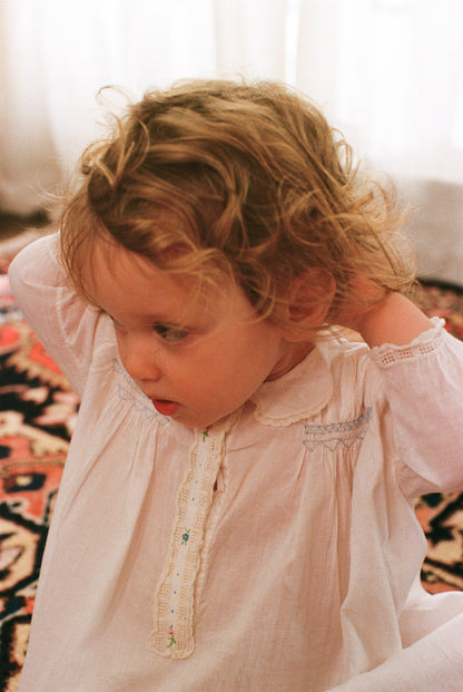 Child wearing a light pink dress with lace details, sitting on a patterned rug.