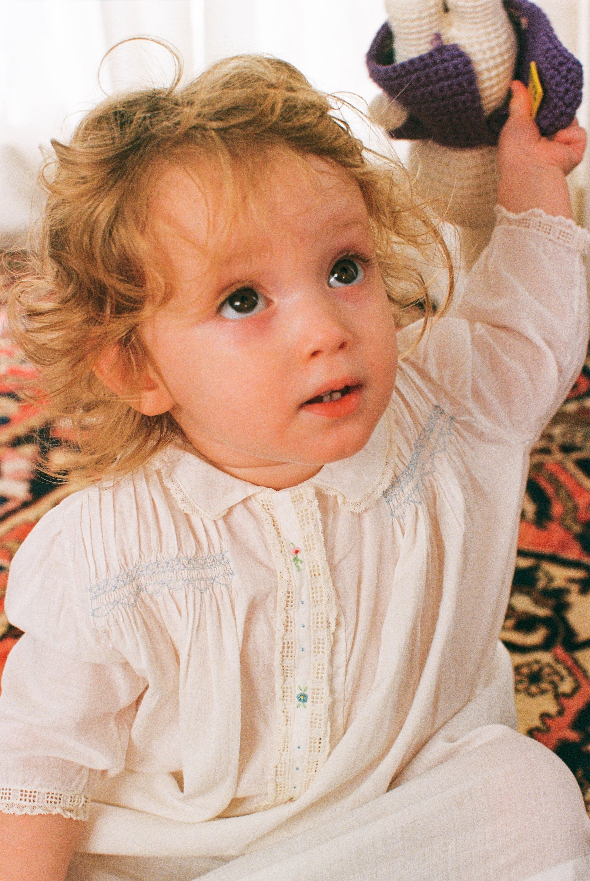 Young child in a white dress holding a stuffed animal against a patterned rug.