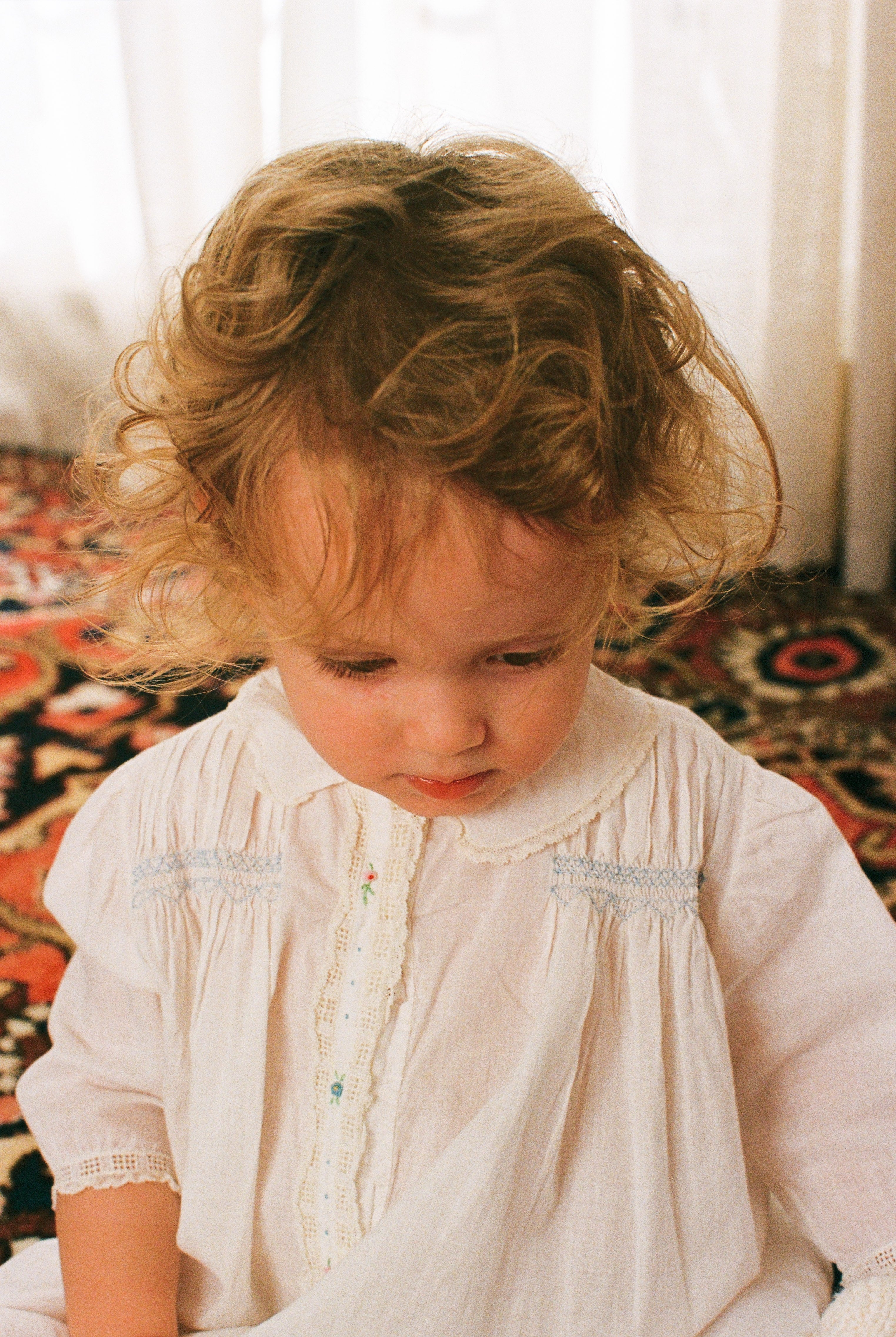 Young child wearing a white dress with lace details, sitting on a patterned rug.