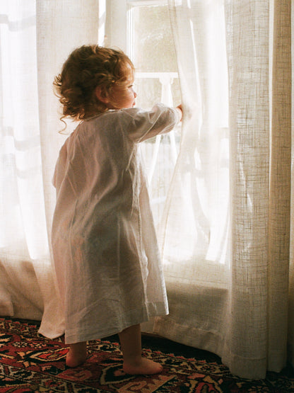Child in a white robe standing by sheer curtains in a softly lit room