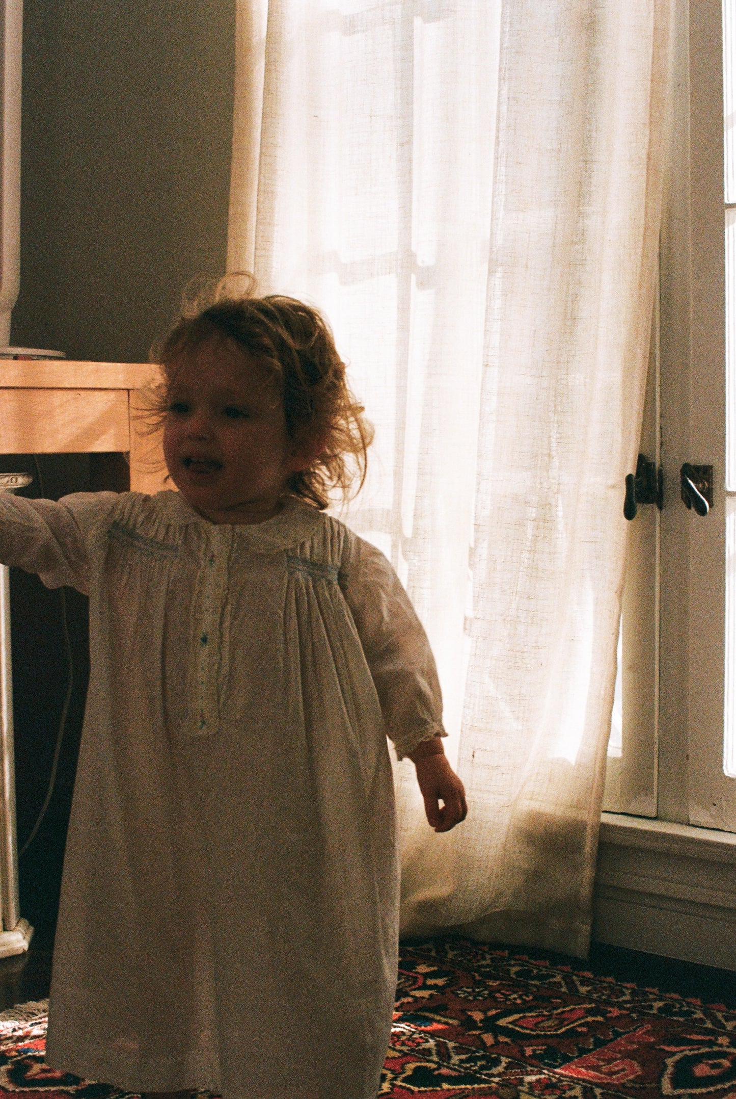 Child in a white dress standing in a room with light filtering through curtains.