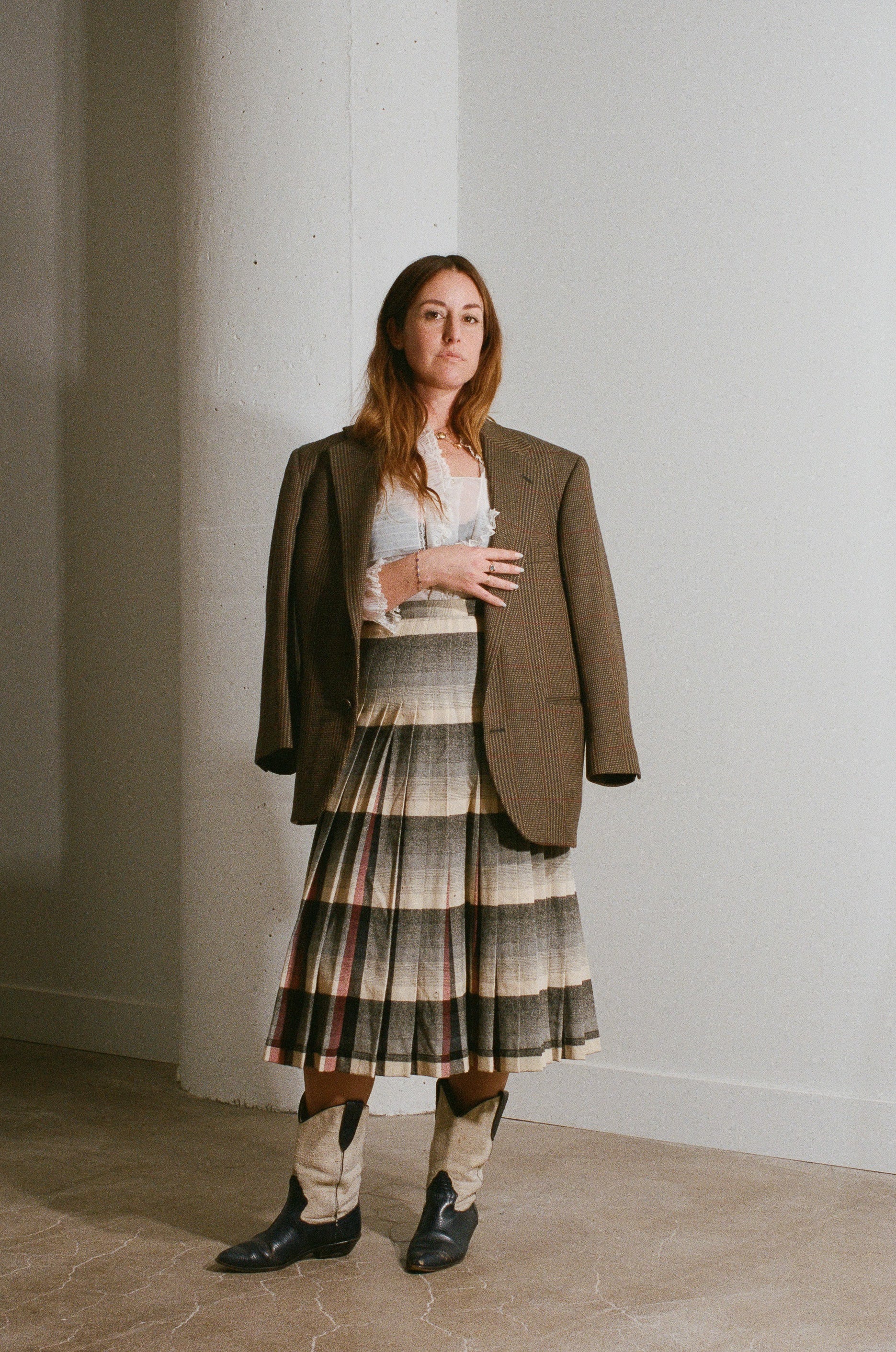 Woman wearing a brown blazer, plaid skirt, and patterned shoes against a white wall.