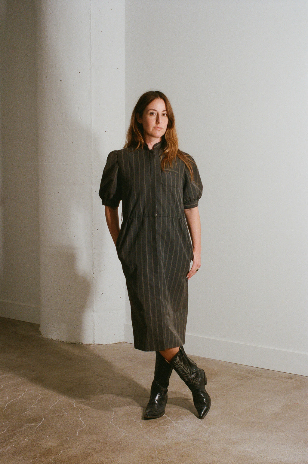 Woman wearing a dark green dress and black boots standing against a white wall.