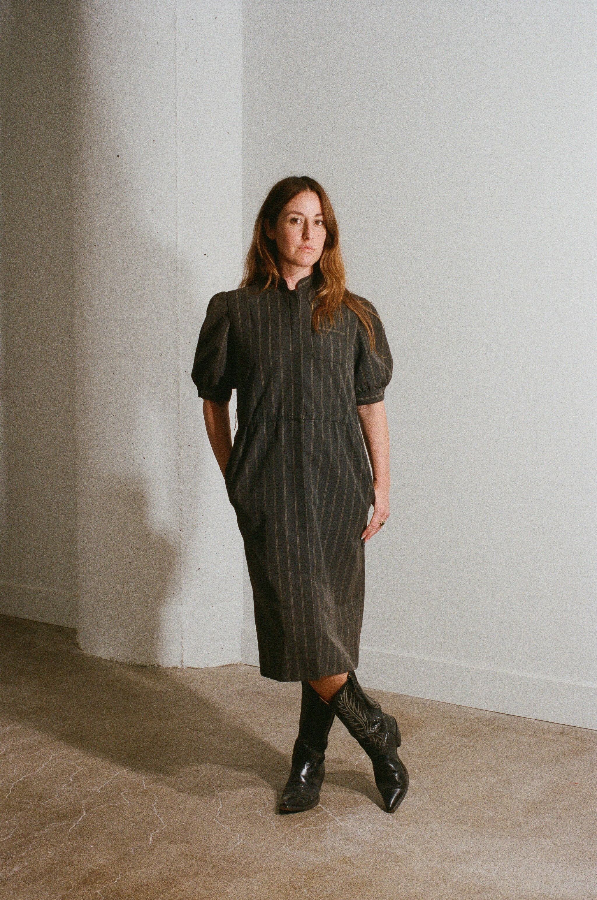 Woman wearing a dark green dress and black boots standing against a white wall.
