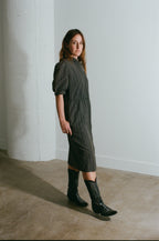 Woman wearing a dark green dress and black boots standing against a white wall.