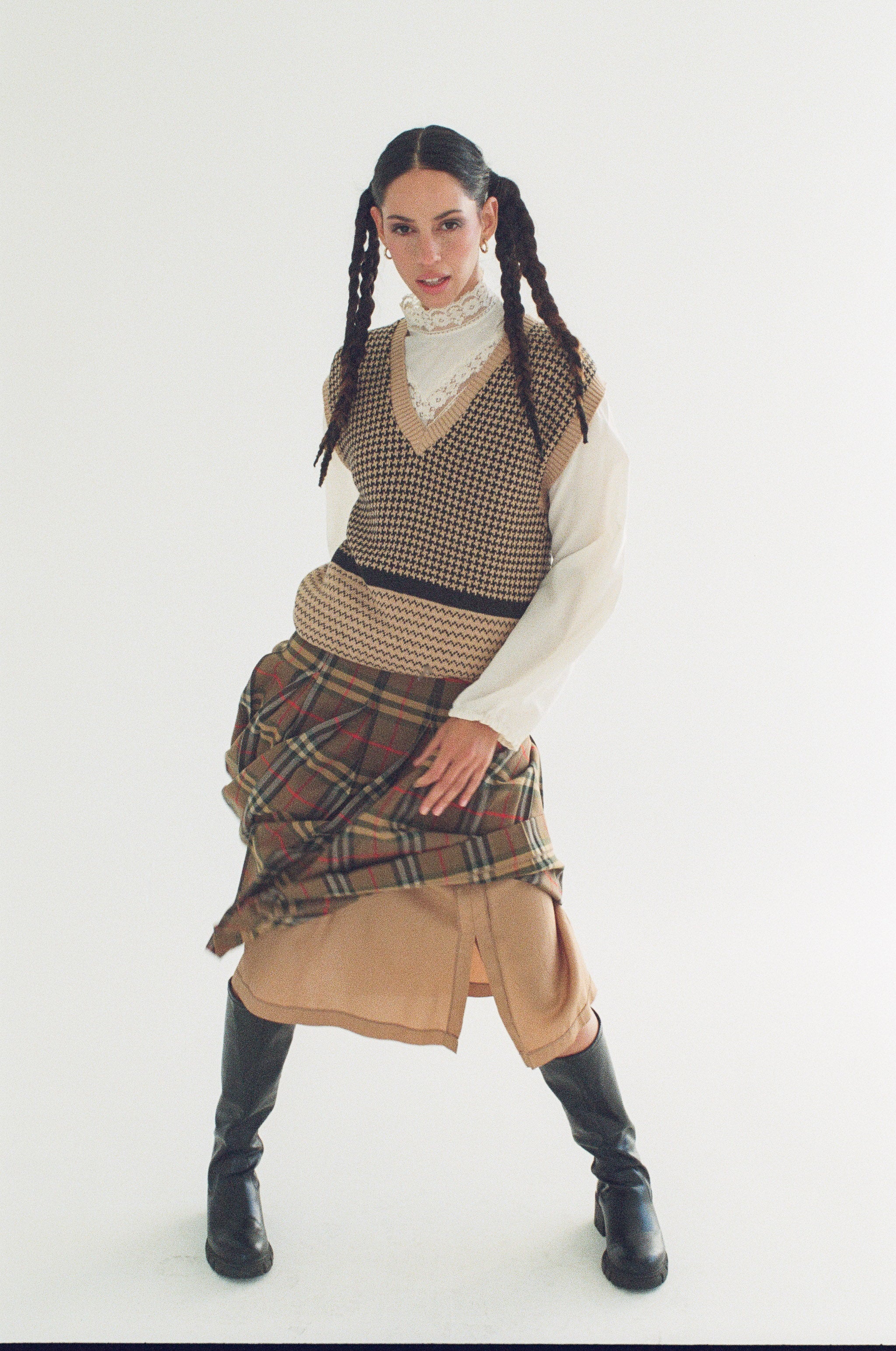 Person wearing a patterned vest, plaid skirt, and beige pants on a white background