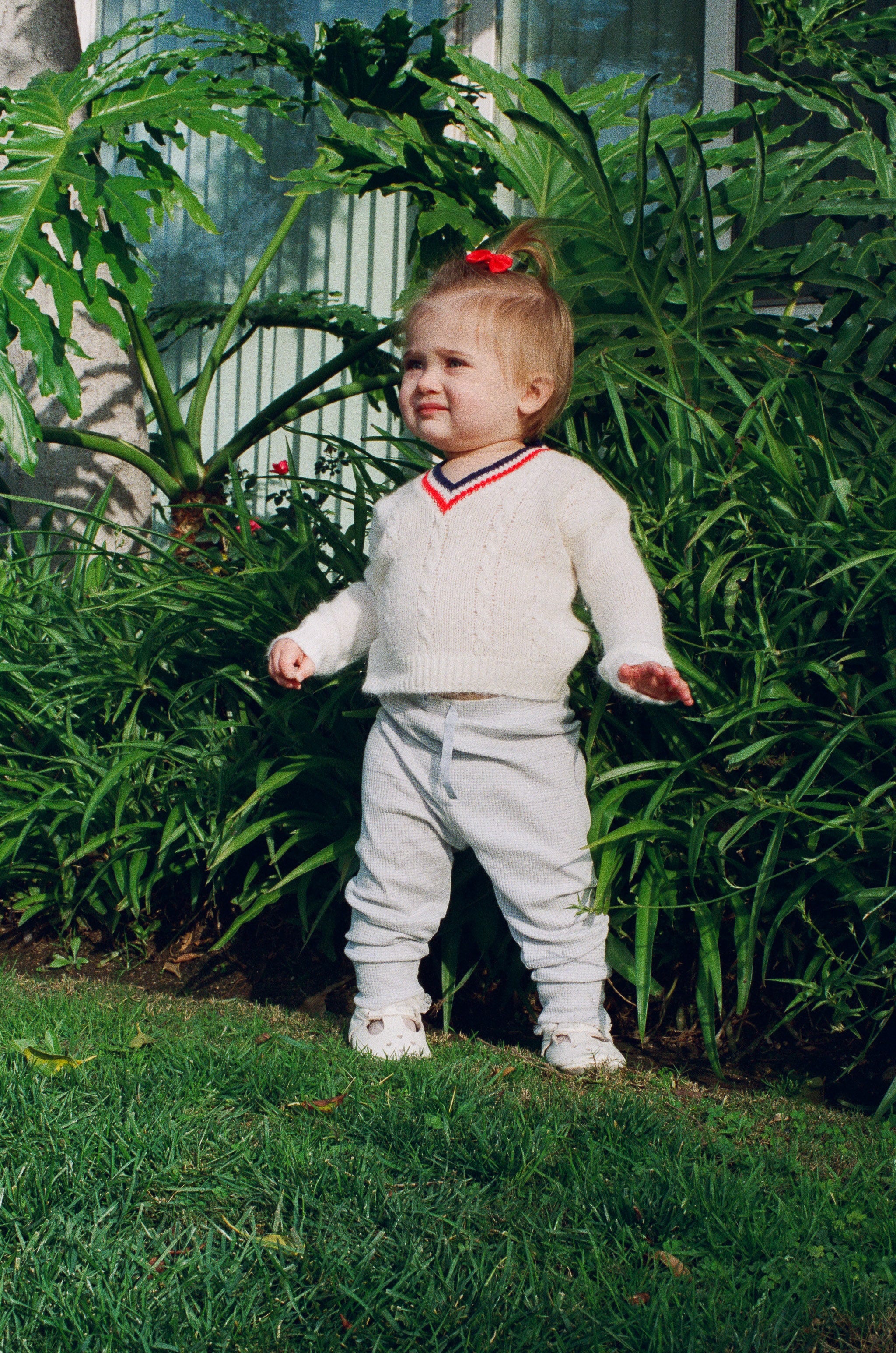 Child in a white sweater and pants standing in a garden with green plants and grass.