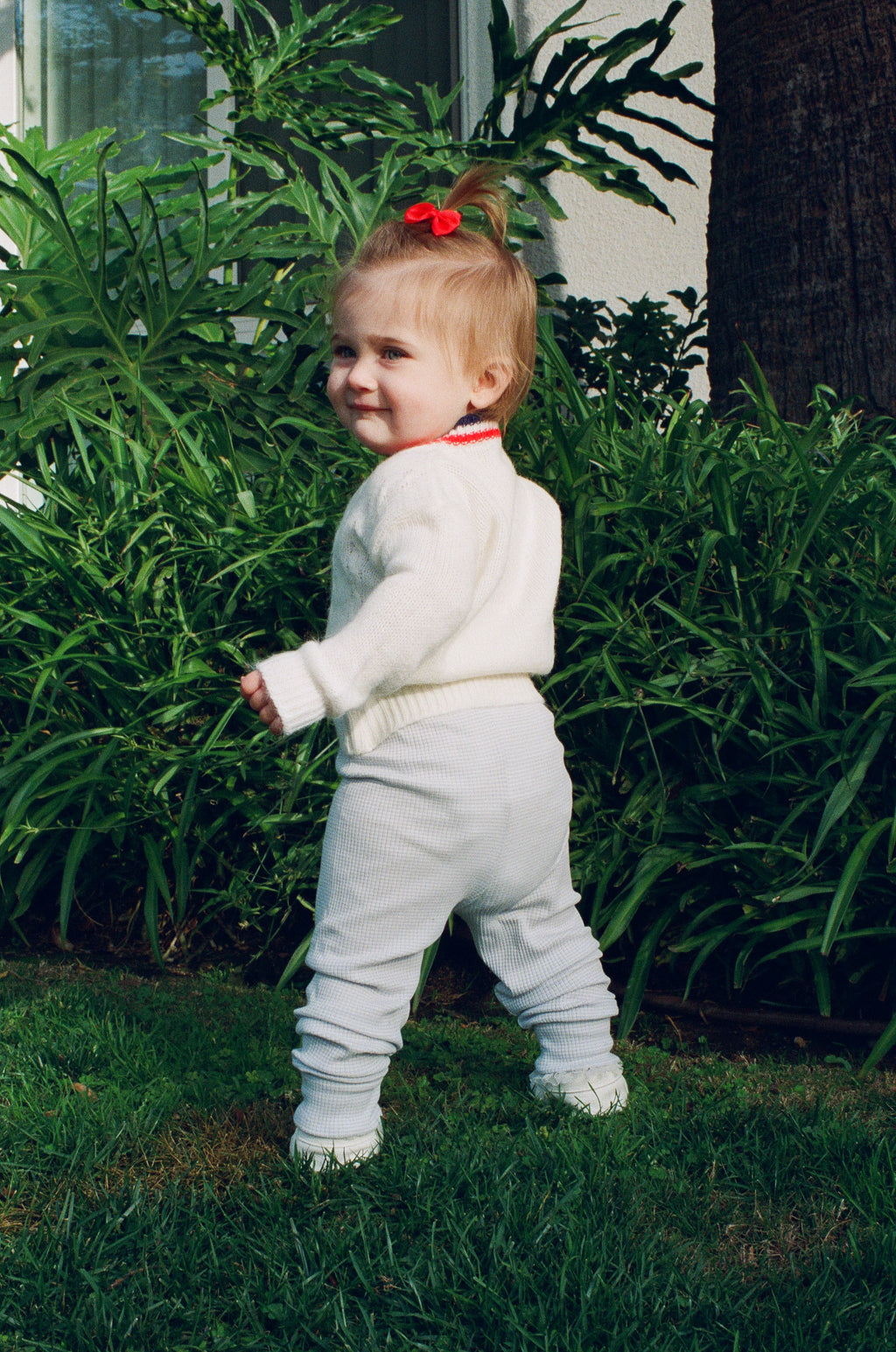 Child in a white outfit standing outdoors with greenery in the background