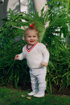 Child wearing a white sweater with red and blue accents, standing in front of green foliage.