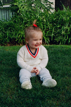 Child sitting on grass wearing a white sweater with a colorful V-neck design.