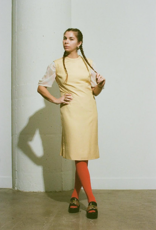 Woman wearing a yellow dress with sheer sleeves against a white wall.