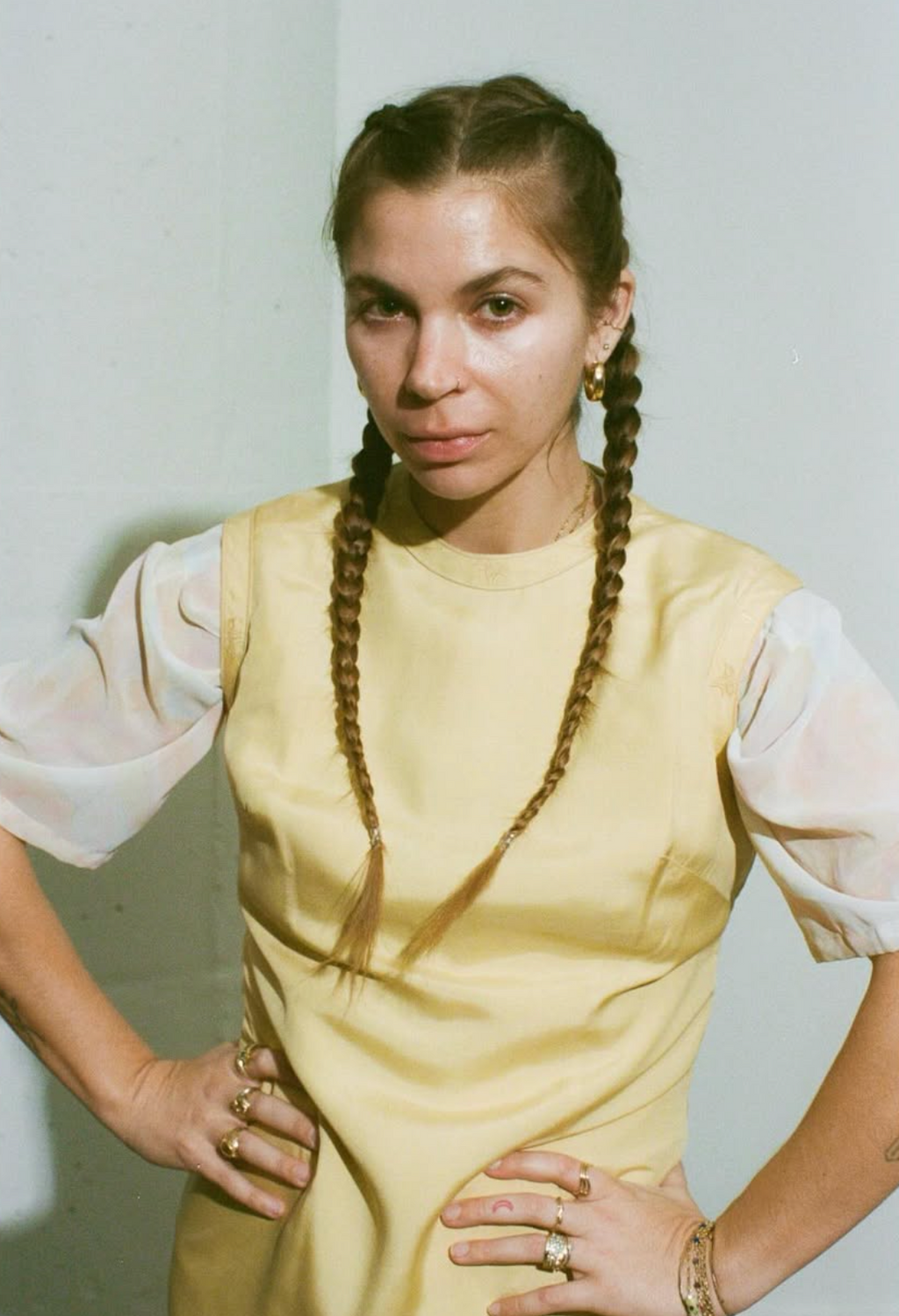 Woman with braided hair wearing a yellow dress and white blouse against a plain background
