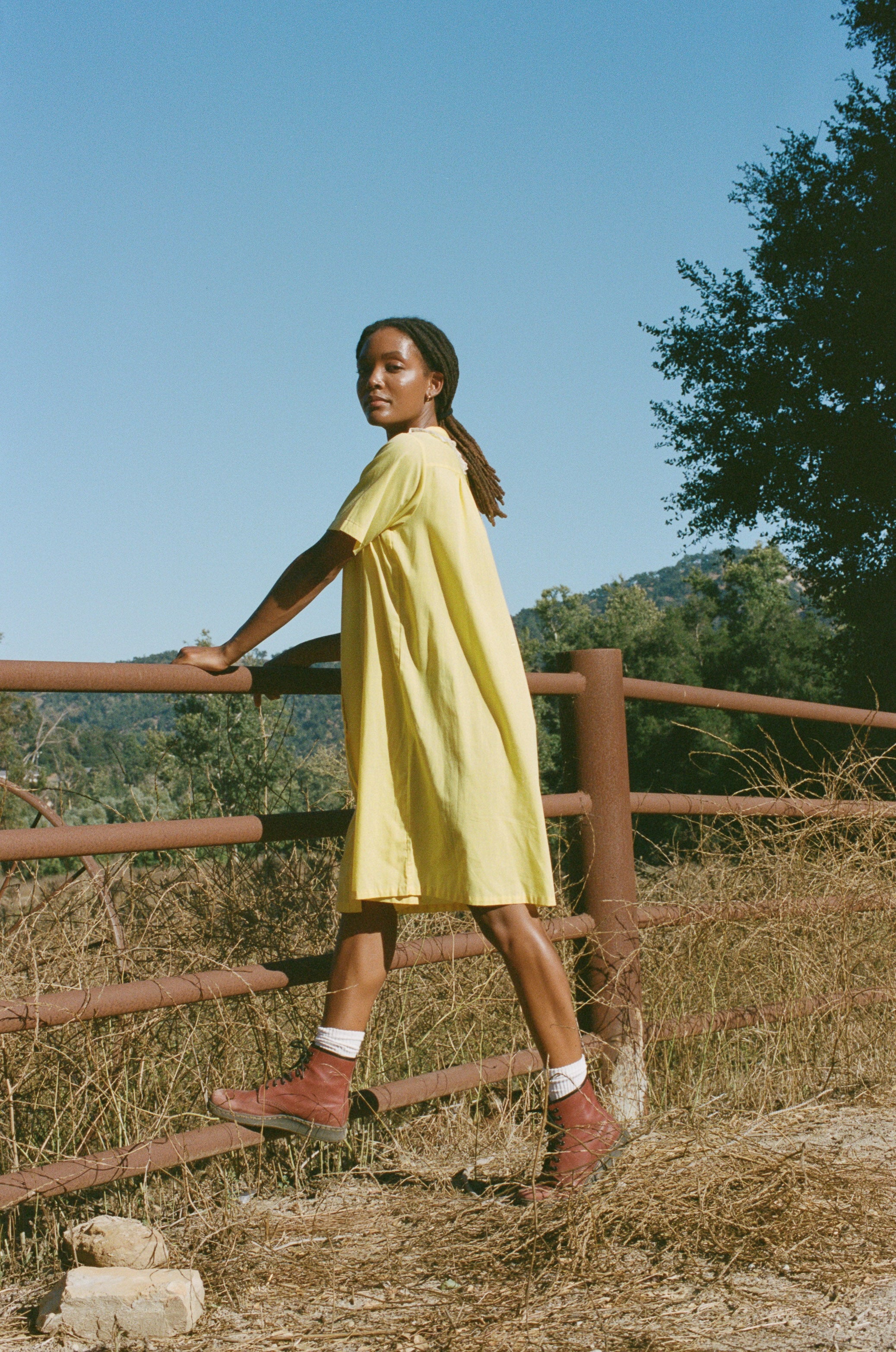 Woman in a yellow dress standing by a wooden fence with a scenic background