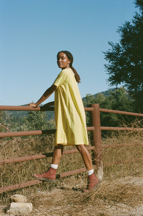 Woman in a yellow dress standing by a wooden fence with a scenic background