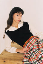 Woman sitting on a wooden floor wearing a black and white top with a plaid skirt.