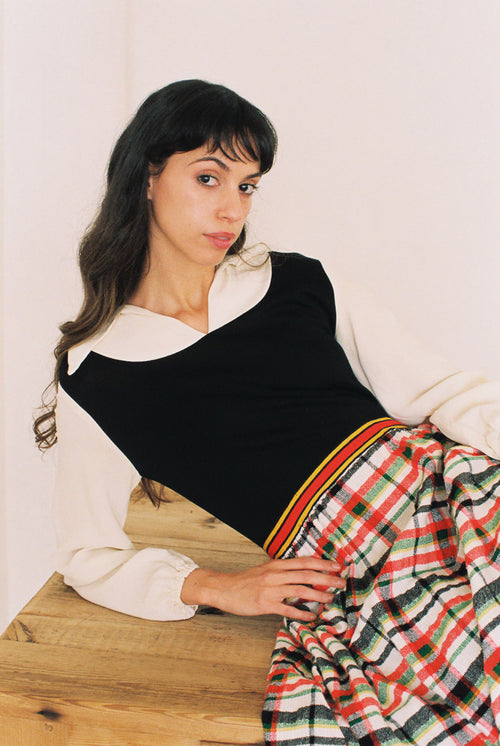 Woman sitting on a wooden floor wearing a black and white top with a plaid skirt.