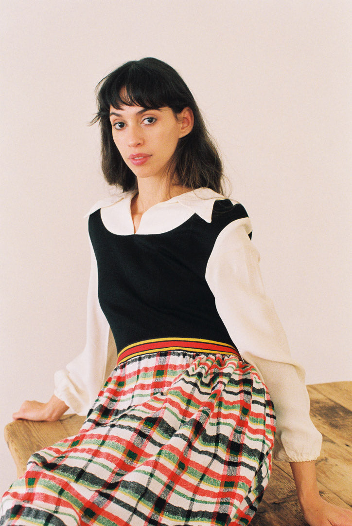 Woman wearing a black top with a white collar and a colorful plaid skirt, sitting on a wooden bench.