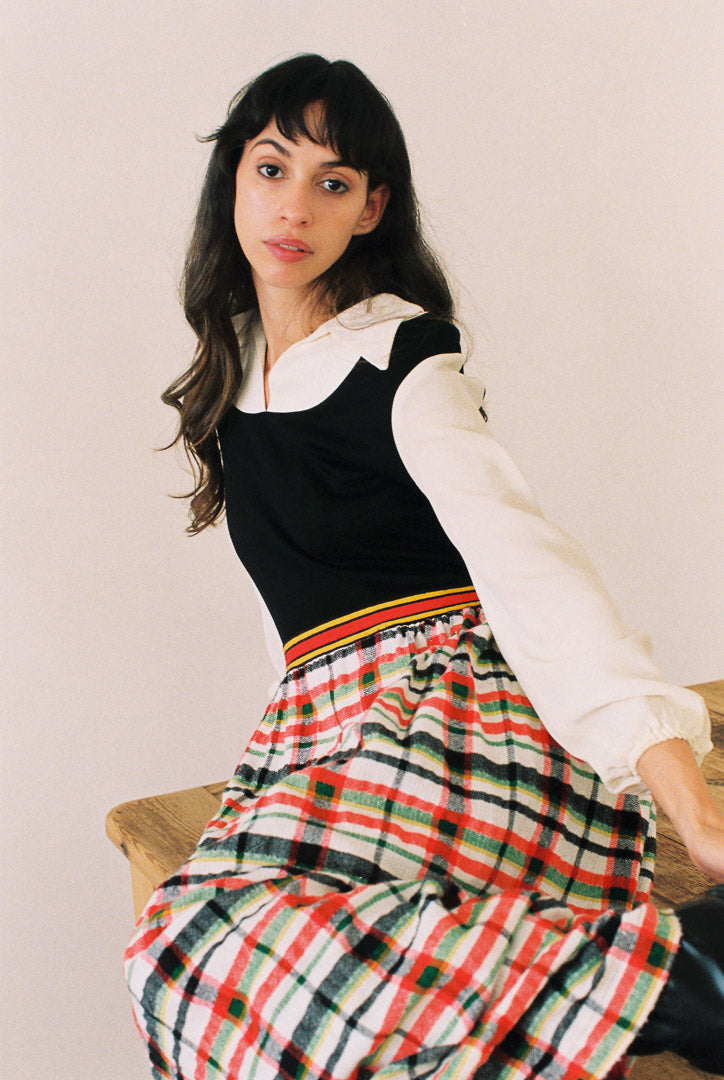 Woman wearing a black top and plaid skirt with a white shirt underneath, sitting on a wooden bench.