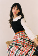 Woman wearing a black top and plaid skirt with a white shirt underneath, sitting on a wooden bench.