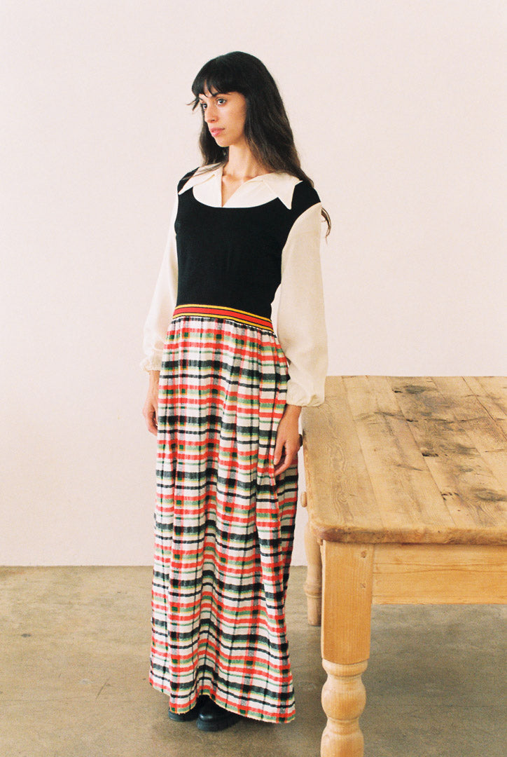 Woman wearing a black top and long plaid skirt standing next to a wooden table.