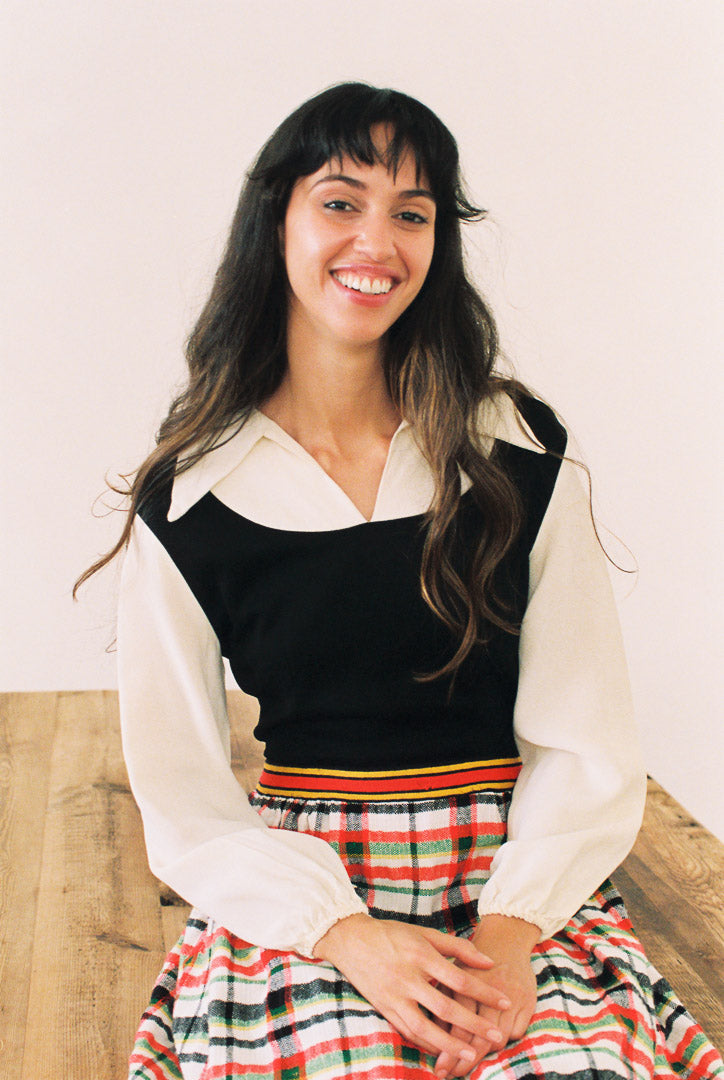 Woman wearing a black vest over a white shirt with a plaid skirt, sitting on a wooden bench.