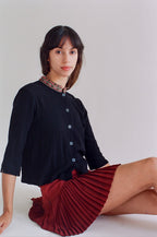 Woman wearing a black top and red pleated skirt sitting on a white surface.