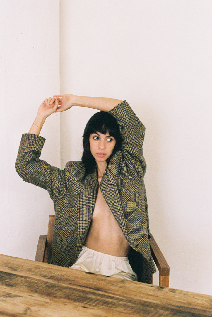 Woman wearing a plaid jacket , sitting on a wooden chair against a white wall.