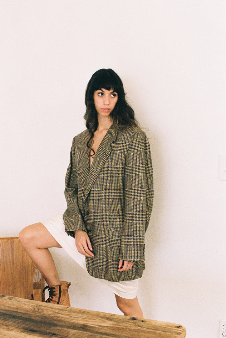 Woman wearing a green checkered blazer over a white top, sitting on a wooden bench against a white wall.