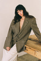 Woman wearing a plaid blazer sitting on a wooden bench against a plain background.