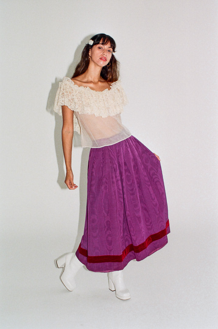 Woman wearing a white top with lace sleeves and a purple skirt on a plain background