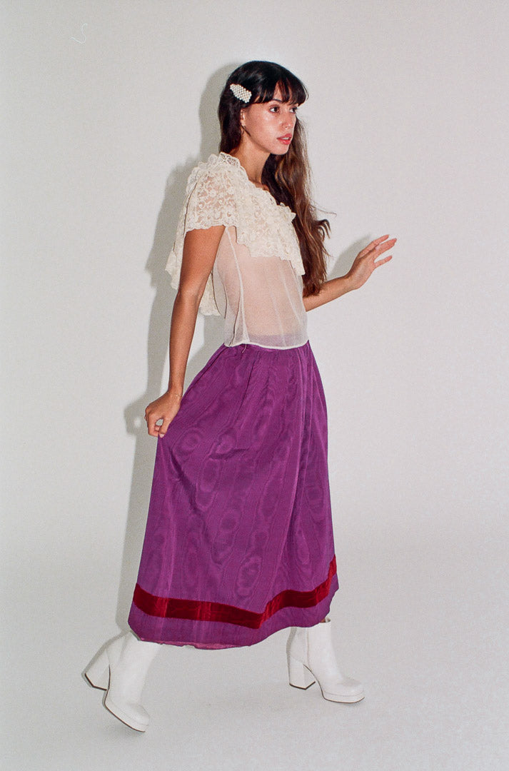 Woman wearing a white lace top and purple skirt with red trim against a white background